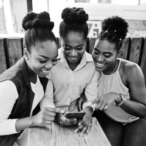 Three african american girls sitting on the table of caffe and looking on mobile phone.